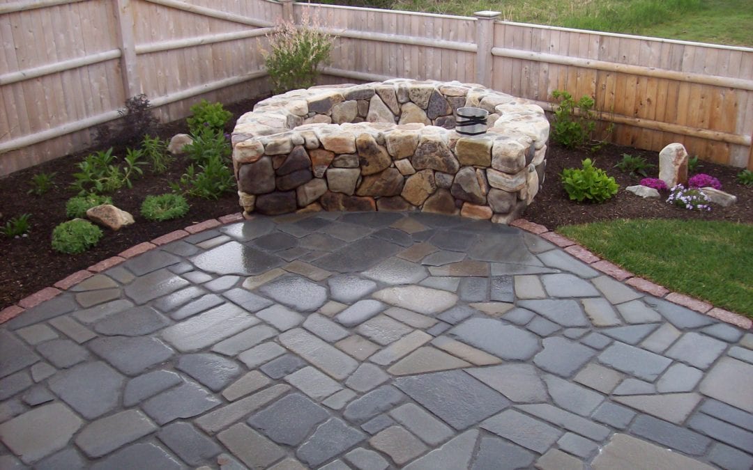 Bluestone patio with natural stone round fireplace surrounded by beautiful landscaping and a wooden fence