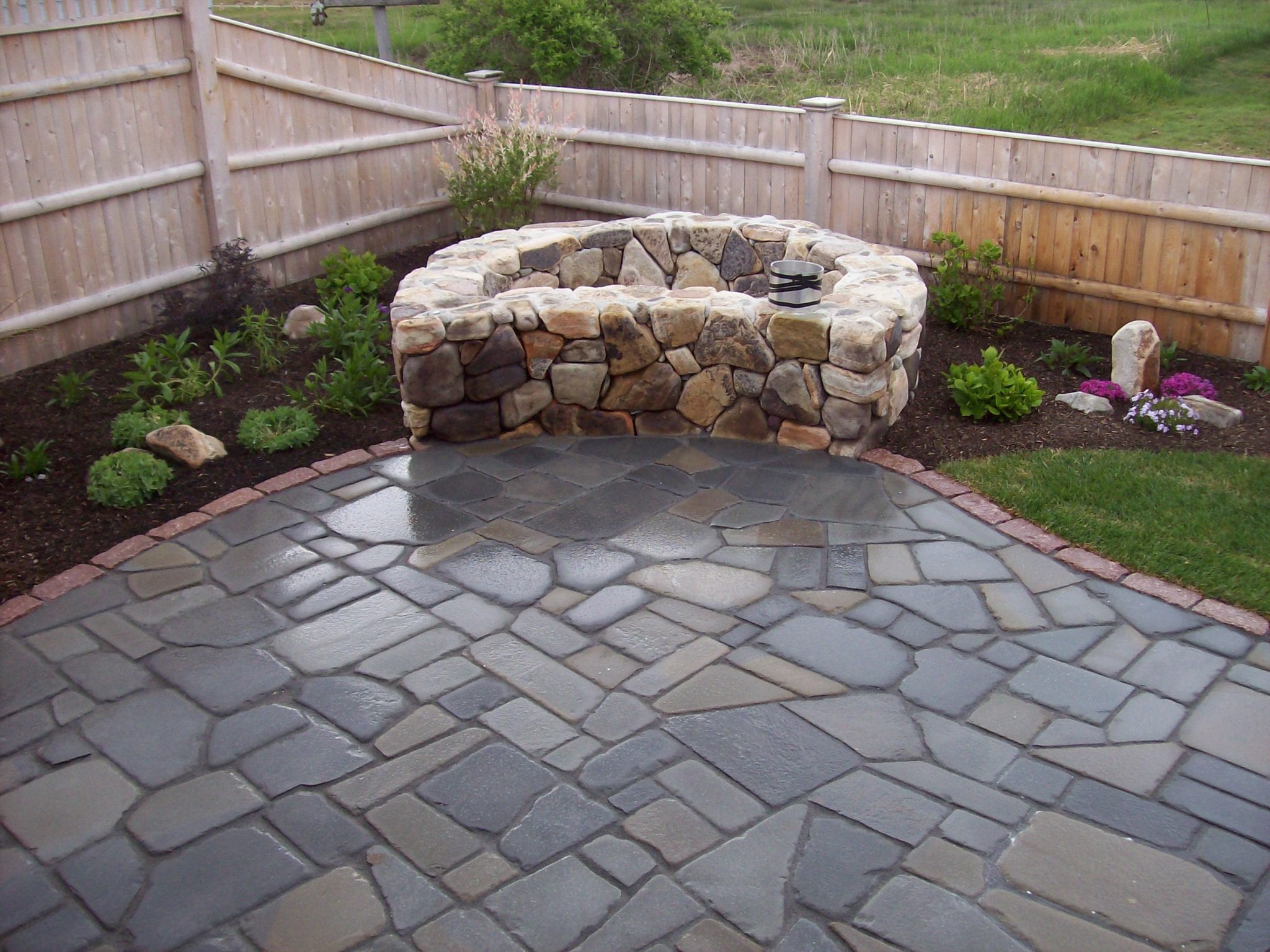 Bluestone patio with natural stone round fireplace surrounded by beautiful landscaping and a wooden fence