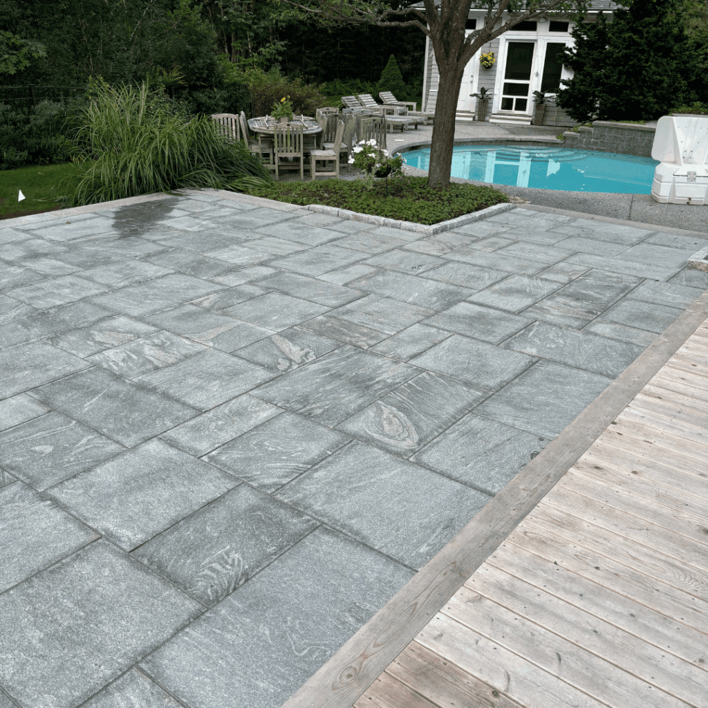 Modern pool patio with granite pavers, outdoor dining area, lounge chairs, and integrated landscape design featuring tree planter island and manicured lawn