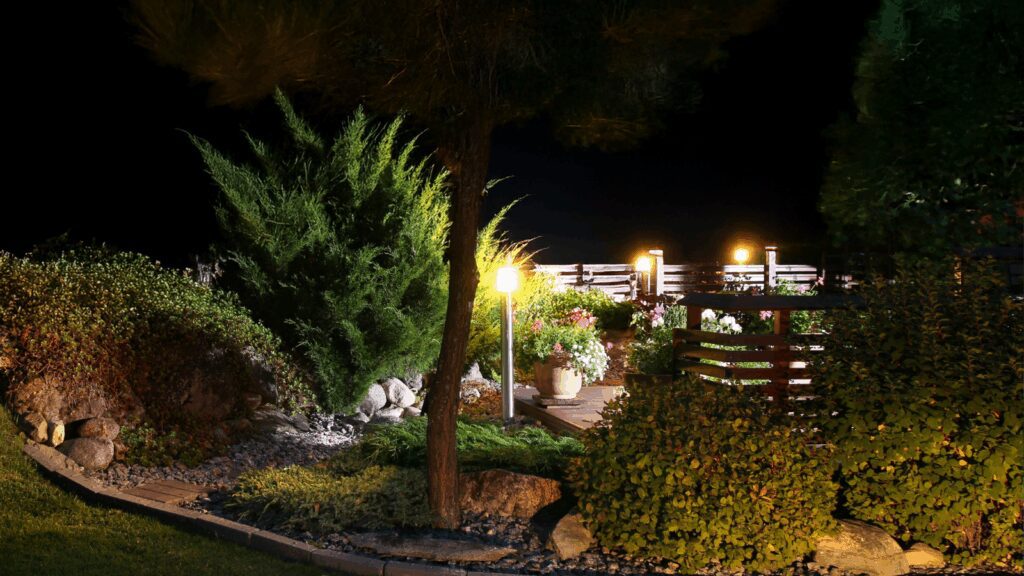 Nighttime garden lighting highlighting wooden deck, evergreen landscaping and decorative plantings, outdoor living space with illuminated pathway and privacy screening