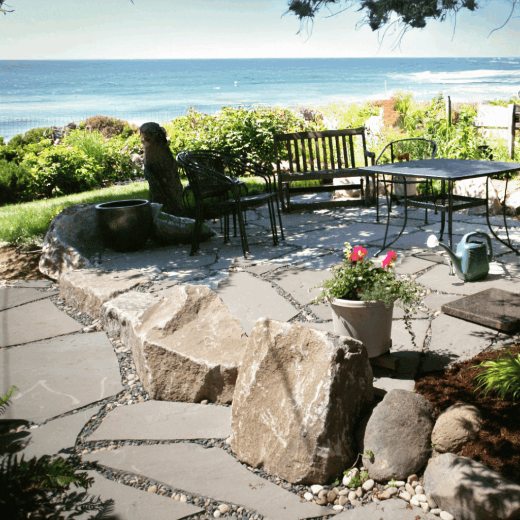 Oceanfront flagstone patio with natural stone elements, wooden bench, and wrought iron furniture offering stunning sea views, coastal garden retreat with potted flowers