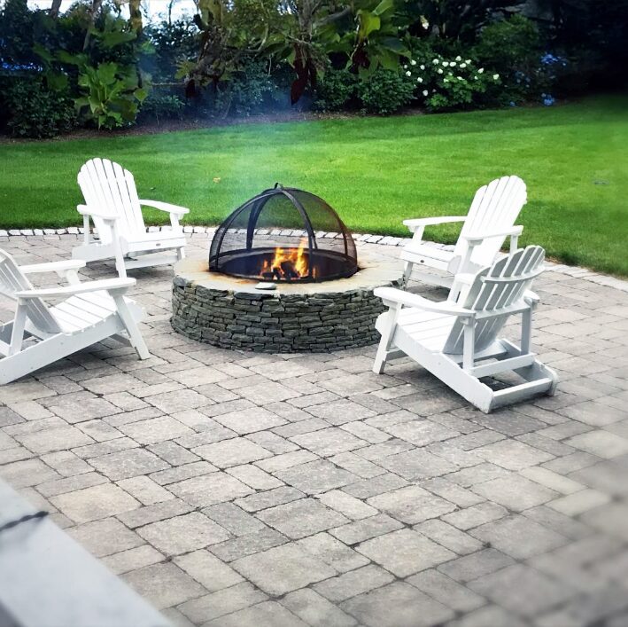 Stone fire pit patio with protective dome screen and white Adirondack chairs, evening outdoor entertainment space surrounded by manicured lawn and garden