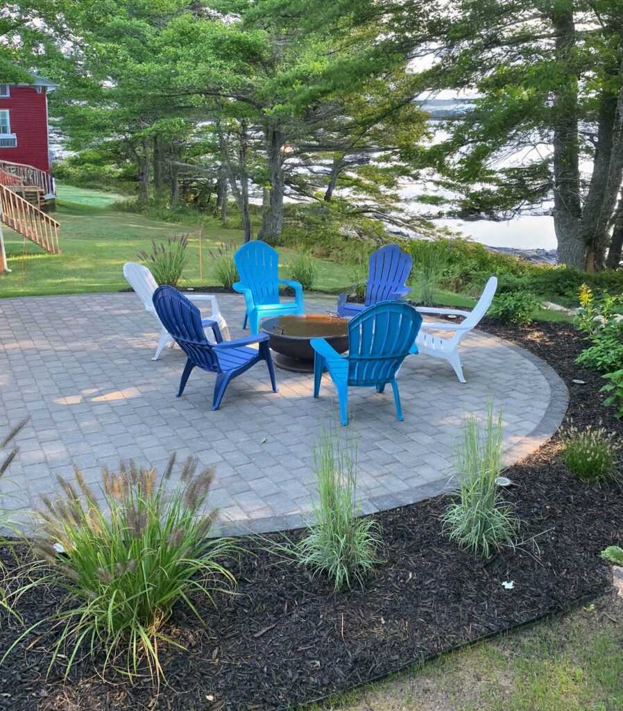 Waterfront paver patio with blue and white Adirondack chairs positioned around metal fire pit, coastal property with view of lake