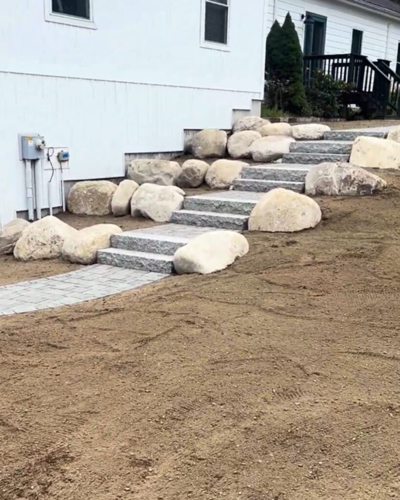 Granite entry steps with a paver walkway winding up a slope beside a white home, lined with natural boulder landscaping in Scarborough, Maine