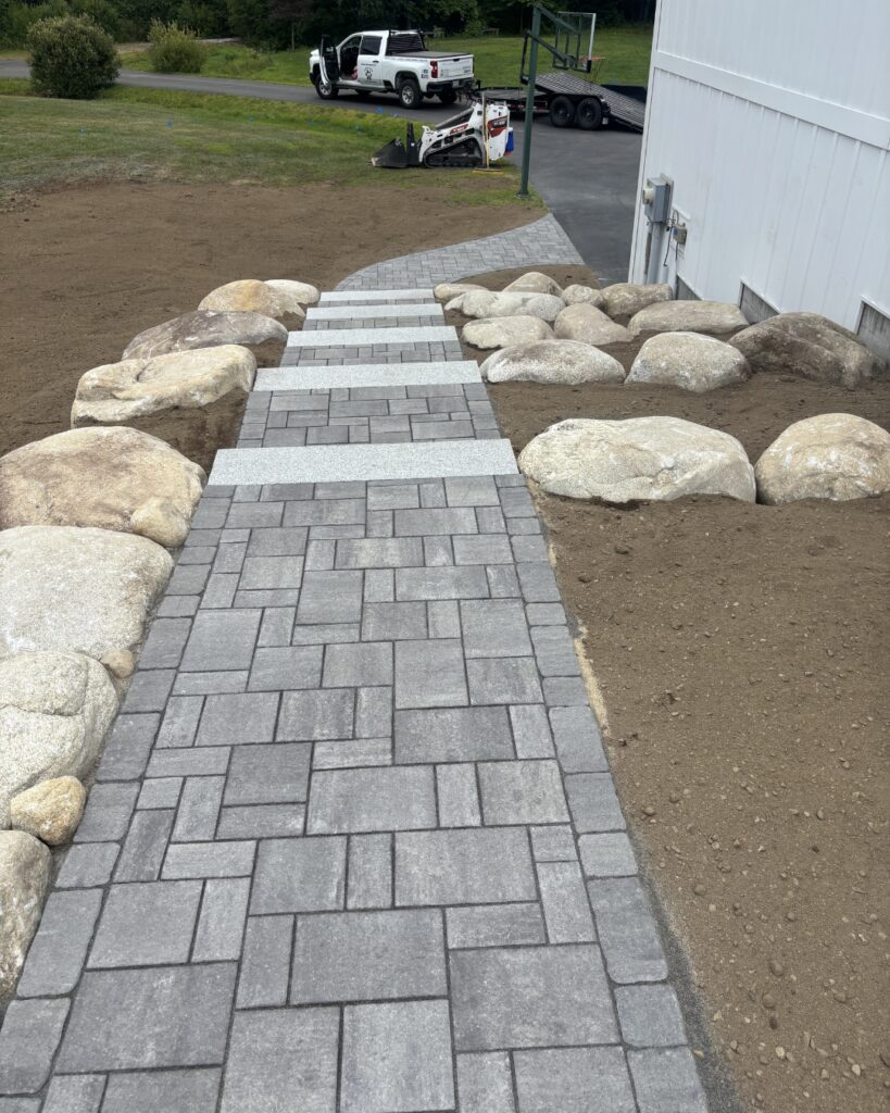 View down a paver walkway with granite step transitions, flanked by large natural boulders set in graded soil, in Scarborough, Maine