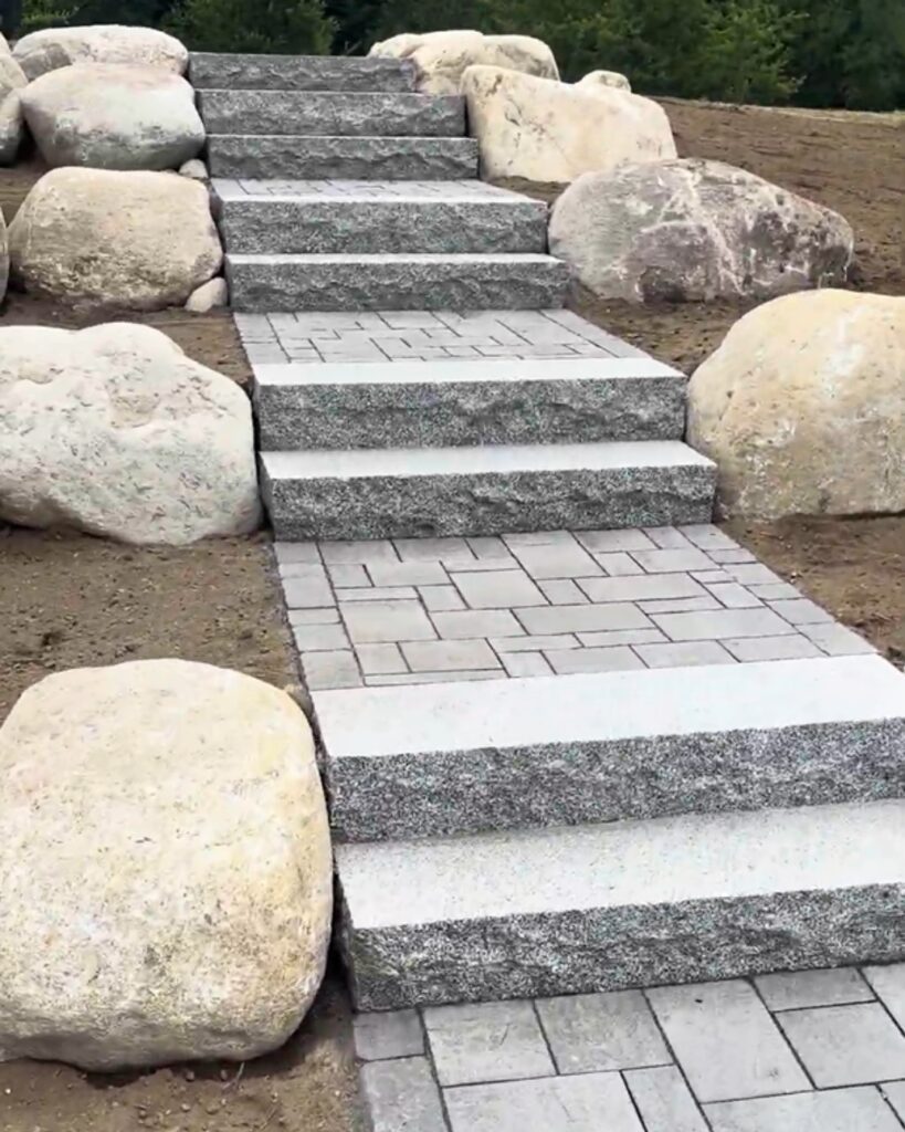 Rough-cut granite steps with Genest Sebago paver landings climbing a hillside, framed by natural boulders, in Scarborough, Maine