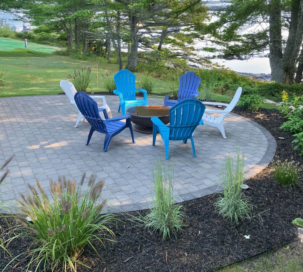 Round paver patio with a fire pit, Adirondack seating, and ornamental grasses near the water in Yarmouth, Maine